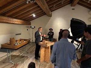 A heritage holder explains a traditional practice beside wooden objects and models while the THRIVE Academy team films the presentation using professional camera and lighting equipment.