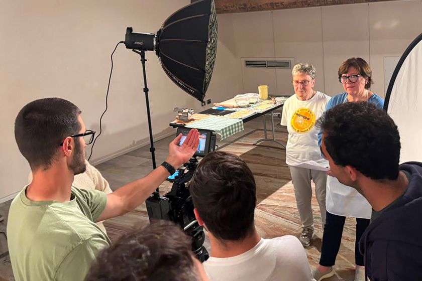Participants at the THRIVE Academy film a heritage demonstration in an indoor workshop space. Two women stand in front of a camera while attendees observe the recording process and learn to use professional lighting equipment, including a studio light and reflector.