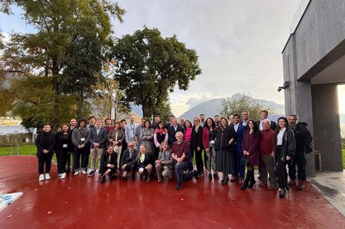 A wide-angle group shot of conference participants posing together outdoors. The setting features a red ground, lush green trees, and a scenic view of mountains and a body of water under a cloudy sky.